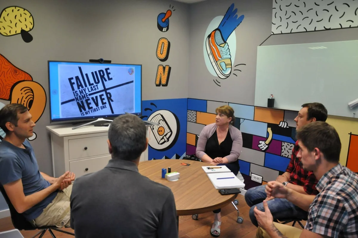 Five participants around a table in a colourful meeting room, with a presentation on a large screen.